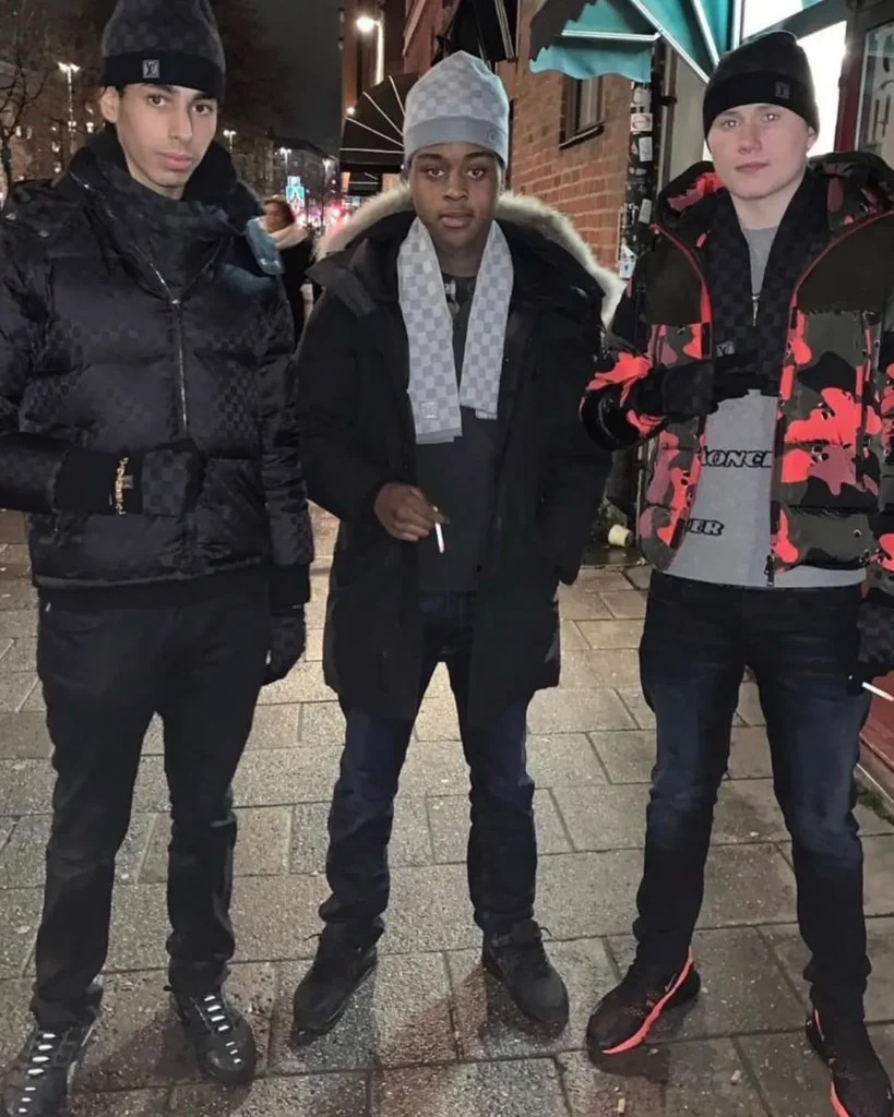Três jovens rappers suecos posam juntos à noite em uma rua urbana de Estocolmo, vestindo roupas de inverno, com looks em preto, cinza e destaque para detalhes em vermelho, evidenciando estilo streetwear típico da cena hip hop escandinava.